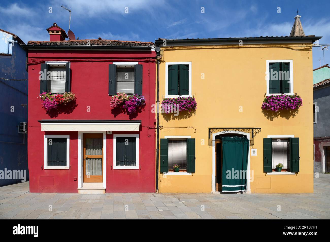 Case rosse e gialle luminose con fiori freschi sulle finestre situate contro il cielo blu nuvoloso sulla strada dell'Isola di Burano a Venezia, Italia Foto Stock