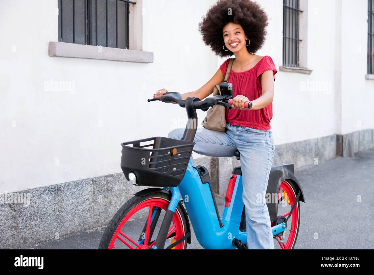 Goditi la giovane donna marocchina con un'acconciatura afro sorridente e guarda la macchina fotografica mentre viaggi in bicicletta elettrica e stai in piedi per strada Foto Stock