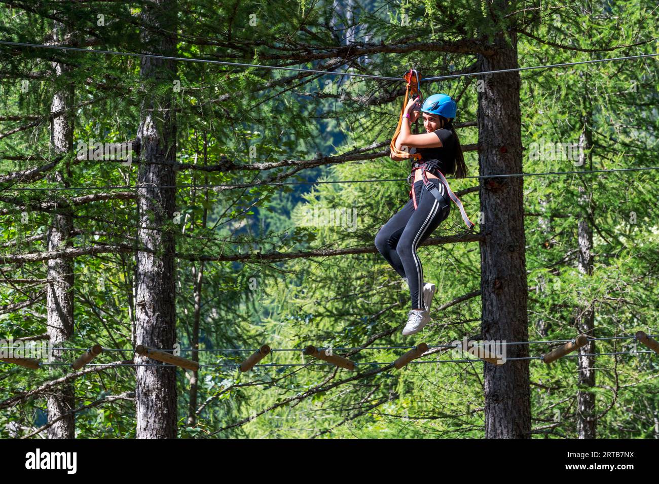 Vista laterale ad angolo ridotto di una giovane donna eccitata nel casco mentre è appesa alla fune superiore con cavi per cintura di sicurezza e cammina sul filo sospeso in Forest adv Foto Stock