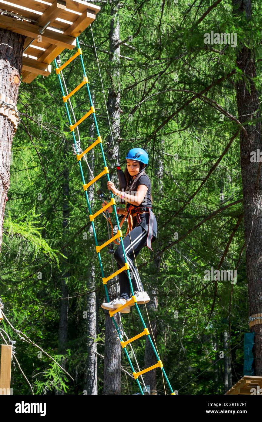 Vista laterale di una giovane donna eccitata con il casco che guarda in basso mentre si arrampica la scala appesa dalla panca di legno con cavi di sicurezza alla luce del sole durante l'avventura Foto Stock
