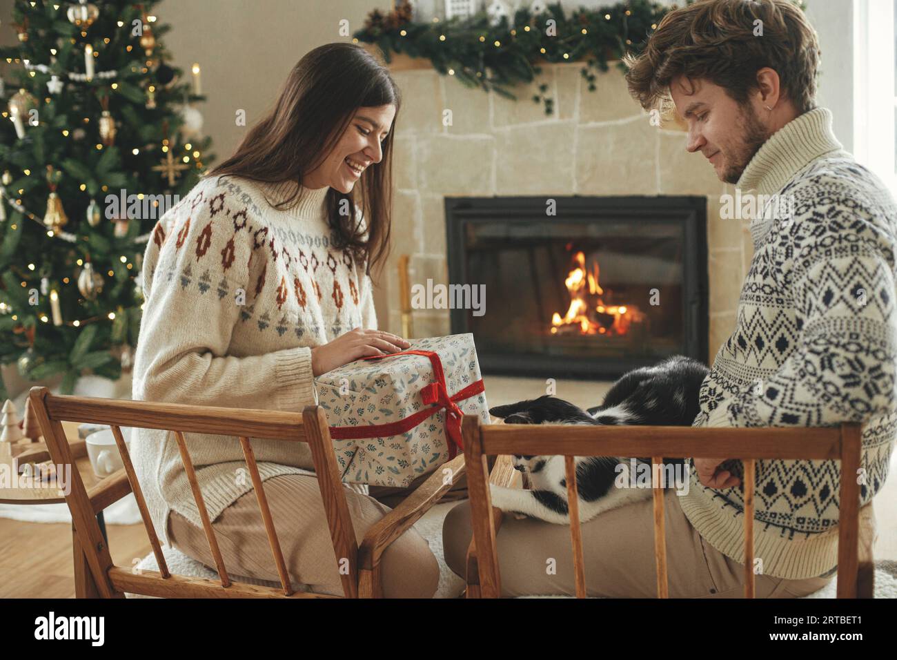 Felice coppia con intimi maglioni che giocano con il gatto e scambiano eleganti regali di natale sullo sfondo del camino con il mantello festivo e il cristo moderno Foto Stock