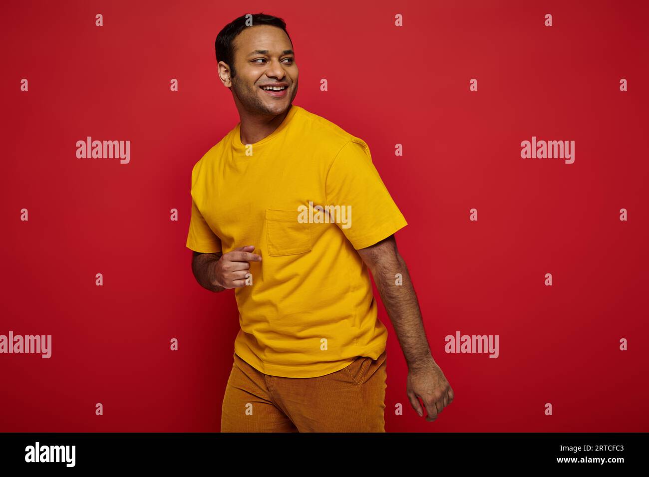 un uomo indiano brillante con abiti casual e luminosi che guardano lontano e sorridono su sfondo rosso, ottimista Foto Stock