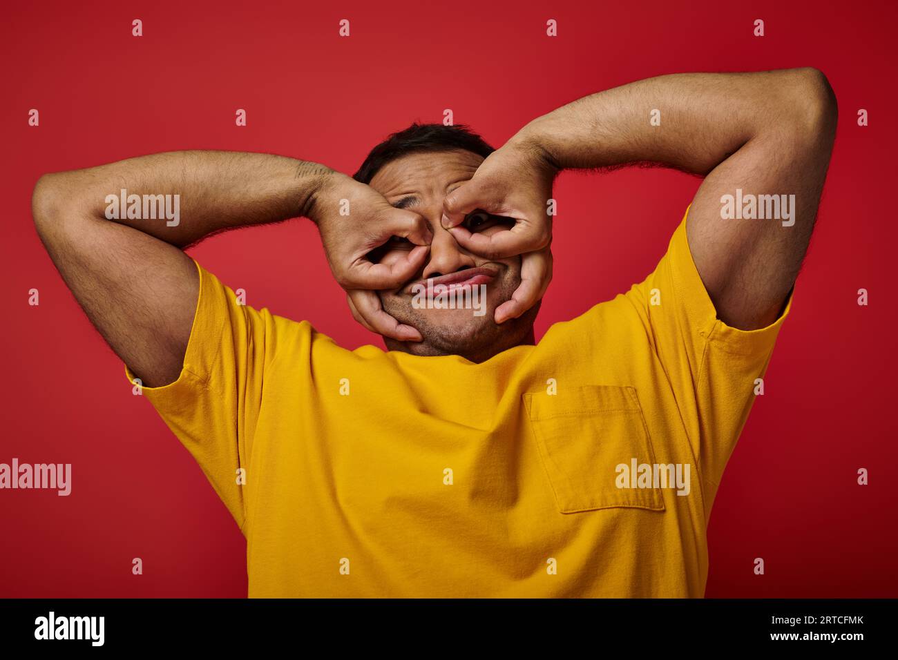 simpatico indiano con t-shirt gialla grigia e gesti su sfondo rosso, faccia espressiva Foto Stock