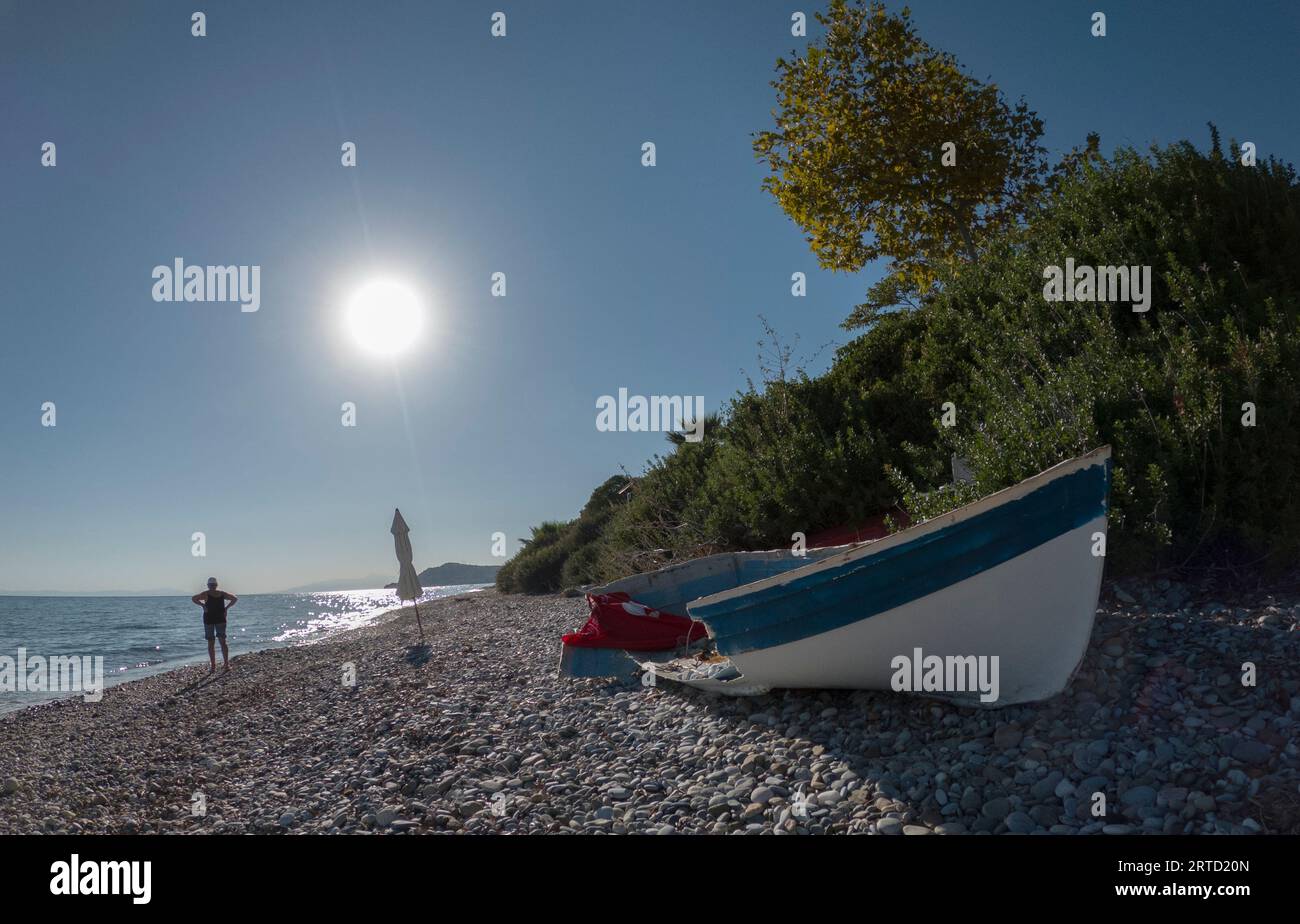 Una barca abbandonata e rotta e la stessa sedia si trova sulla riva del mare, circondata da cespugli e alberi con una casa in lontananza in Grecia Foto Stock