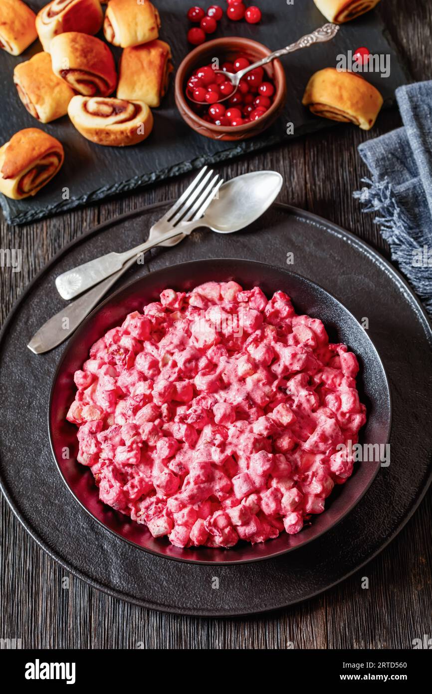 Insalata cremosa di mirtilli rossi con mini marshmallow, mela fresca e noci pecan in ciotola nera su tavolo di legno scuro con rotoli alla cannella, ricetta di Natale, ve Foto Stock