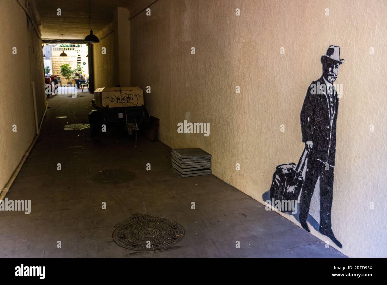 Uno stencil di un uomo con un cappello e una valigia può essere visto su una parete in un passaggio. Sullo sfondo c'è un'area all'aperto con una persona e uno scooter. Tbilisi, Georgia, Bombora Street Foto Stock