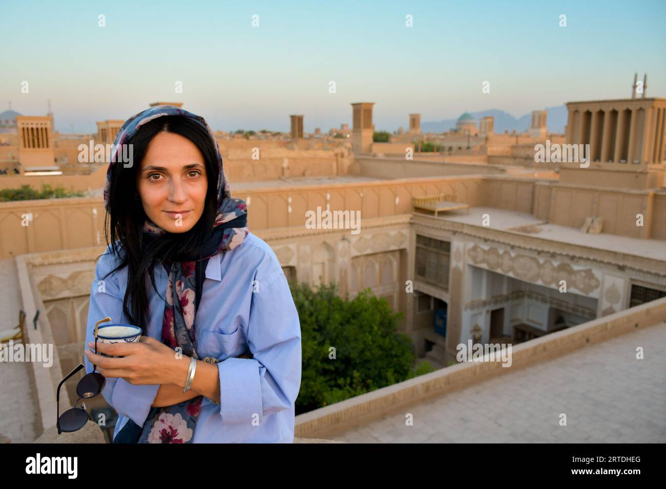 Yazd, Iran - 19 giugno 2022: Un'occhiata turistica caucasica femminile in posa per la macchina fotografica mentre bevi caffè persiano nel tradizionale caffè sul tetto, rilassati e goditi una vista panoramica Foto Stock