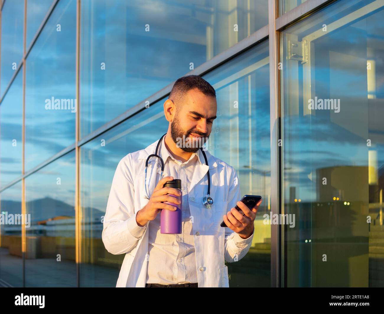 giovane medico maschio che controlla il suo telefono all'aperto di fronte a un ospedale con un caffè thermos sulla mano Foto Stock