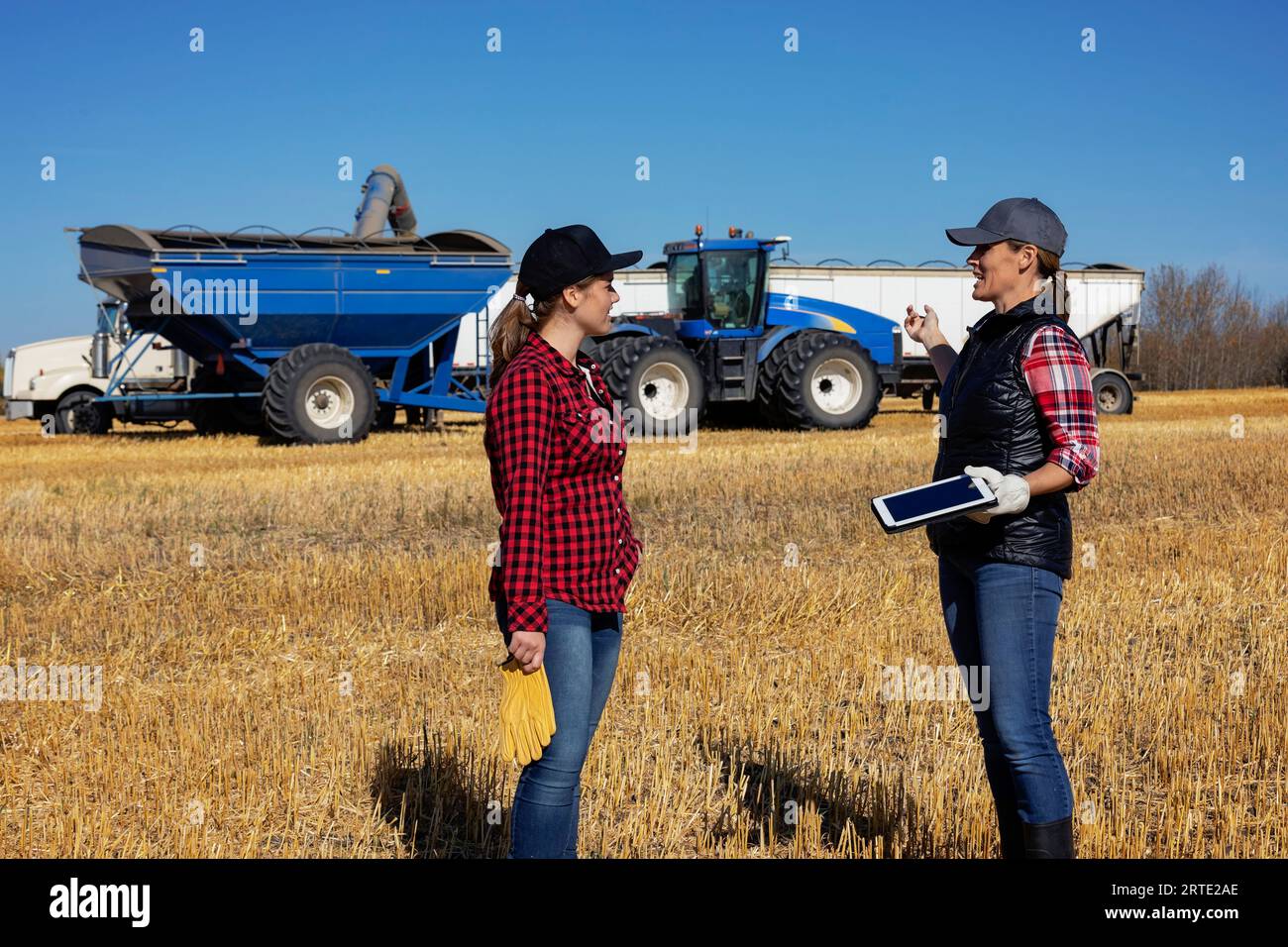 Una donna di fattoria matura in piedi in un campo che insegna al suo apprendista, una donna più giovane, che utilizza tecnologie agricole avanzate durante un raccolto di grano in fa... Foto Stock