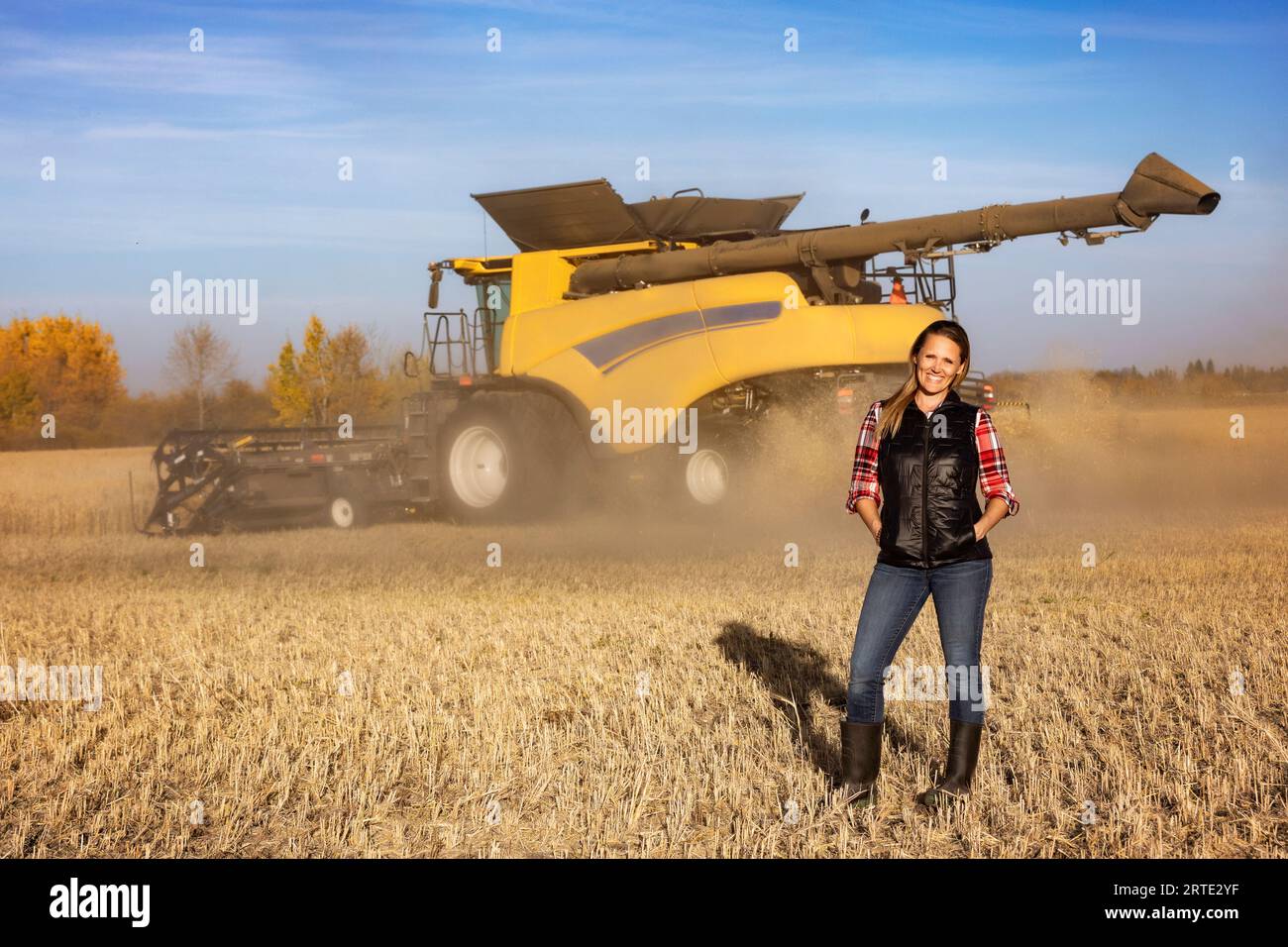 Ritratto di una donna di fattoria matura in piedi in un campo di grano, in posa per la macchina fotografica durante il raccolto con una mietitrebbia che lavora sullo sfondo al tramonto Foto Stock