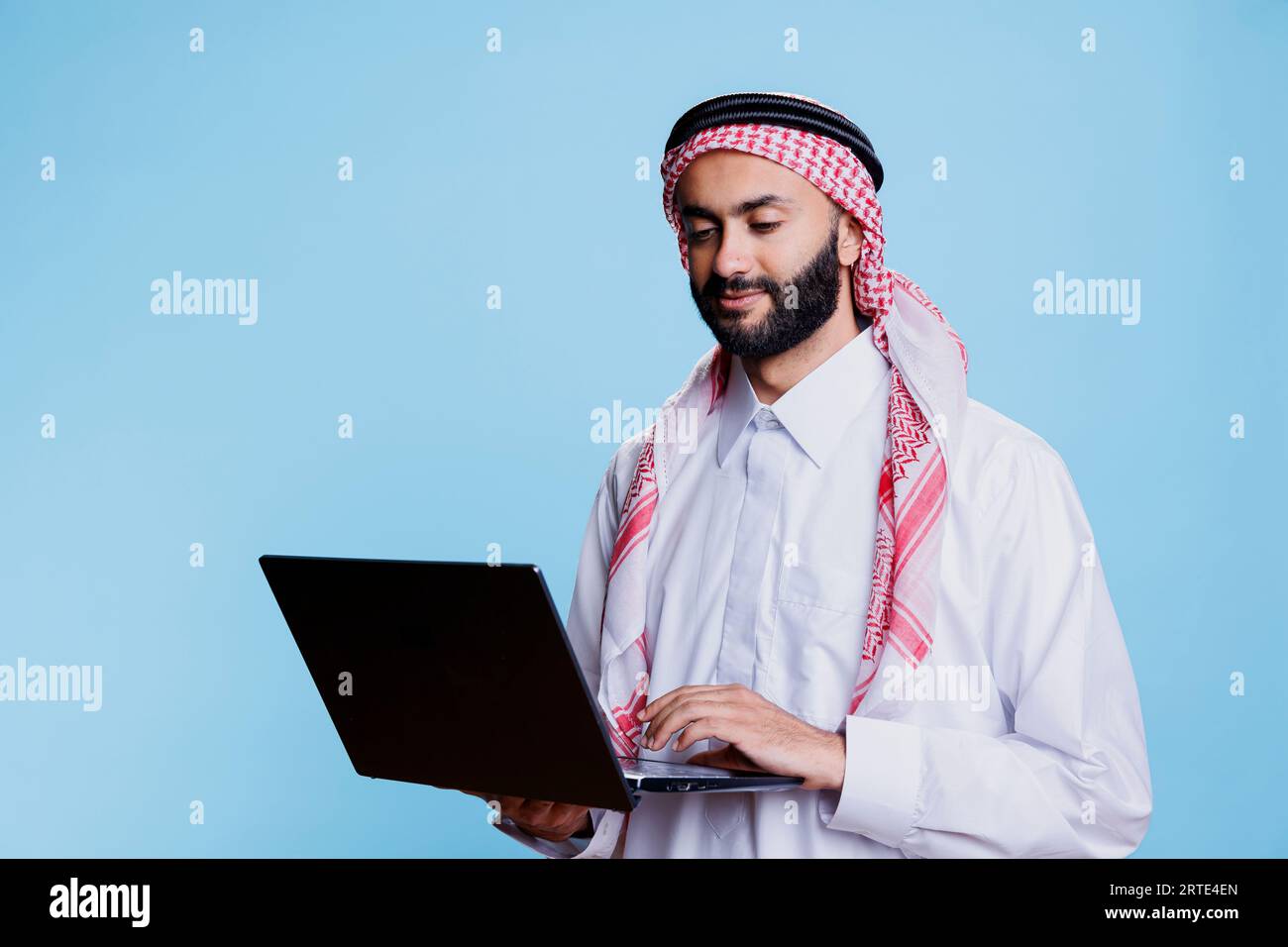 Uomo musulmano che indossa una stoffa tradizionale e un foulard ghutra che regge un laptop e un social media. Persona araba vestita con abiti islamici usando un computer portatile e controllando la pagina Internet Foto Stock