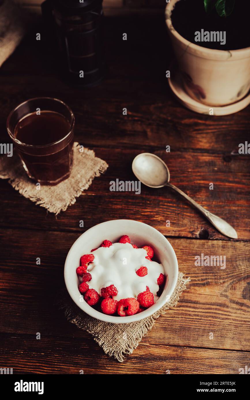 Ciotola di dessert salutare con lamponi maturi e yogurt e bicchiere di caffè su superficie di legno Foto Stock