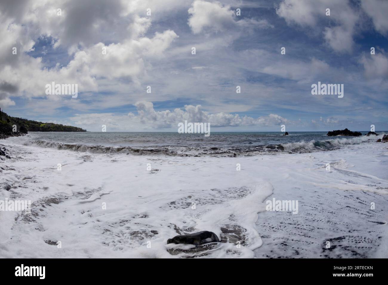 Vista panoramica dell'Oceano Pacifico con il surf schiumoso che copre la riva della spiaggia di sabbia lavica nera sotto un cielo blu e nuvoloso lungo la strada per Hana, s... Foto Stock