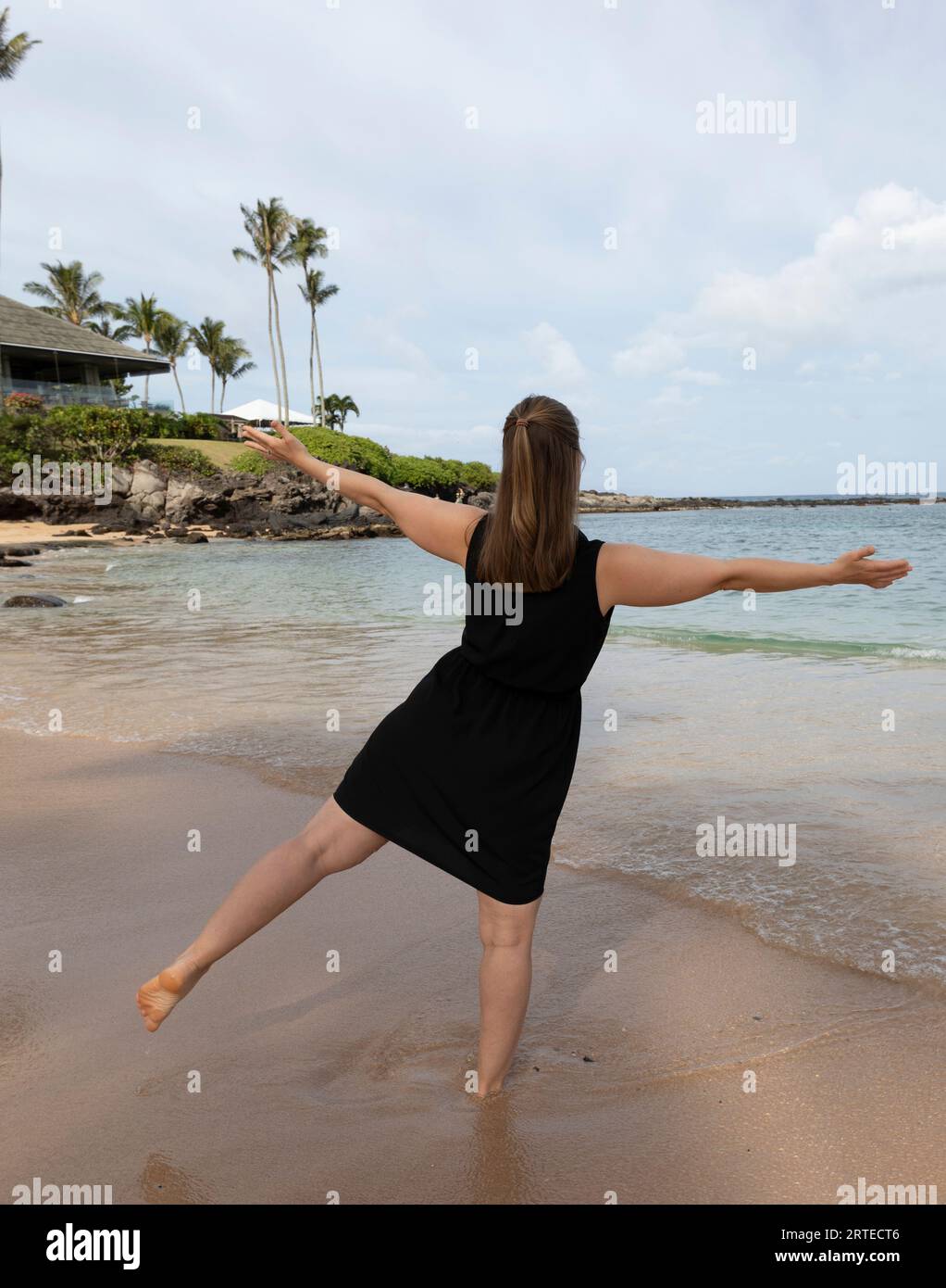 Vista da dietro di una donna in piedi su una gamba nella sabbia bagnata lungo il bordo dell'acqua della spiaggia sabbiosa del Kapalua Resort a West Maui Foto Stock