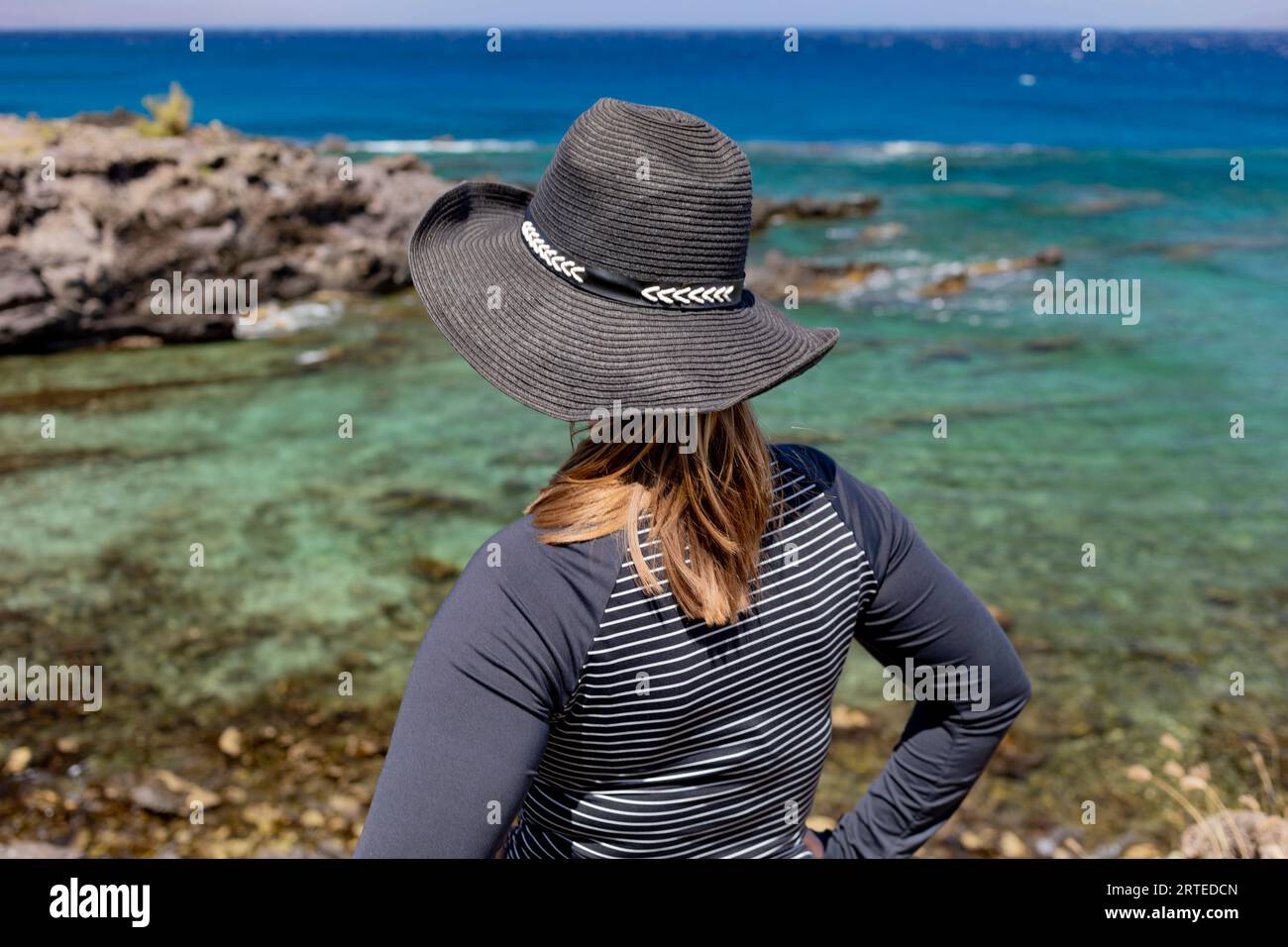 Vista ravvicinata da dietro di una donna che indossa un cappello da sole in piedi sulla riva guardando le acque turchesi sul fronte oceano di Kaanapali Bea... Foto Stock