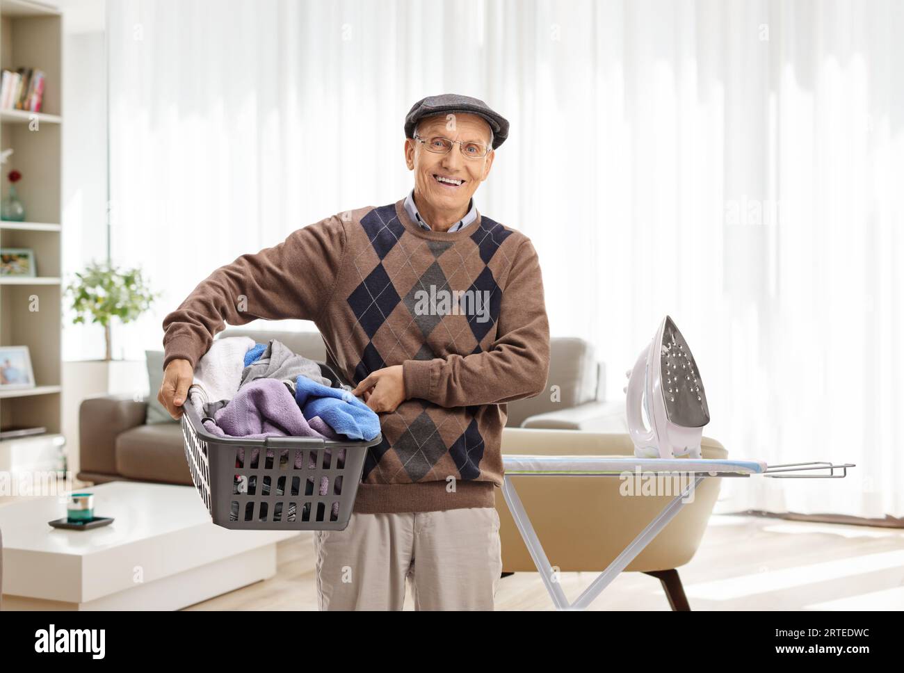 Uomo anziano che tiene un cestino pieno di vestiti davanti a un'asse da stiro in un soggiorno Foto Stock