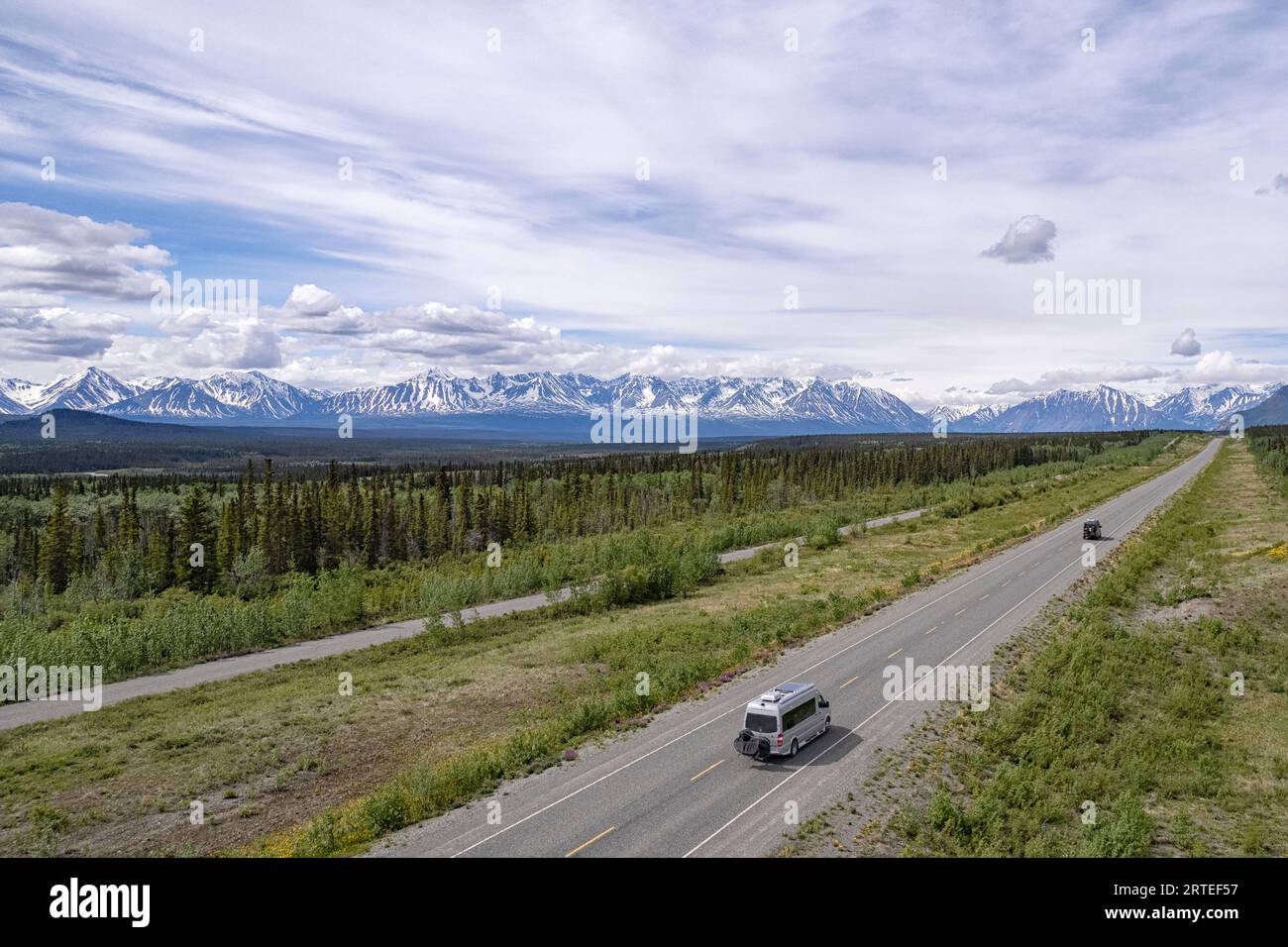 Vista aerea di alcuni camper che viaggiano attraverso il paesaggio boschivo lungo l'Alaska Highway, con fantastiche vedute delle catene montuose innevate su... Foto Stock