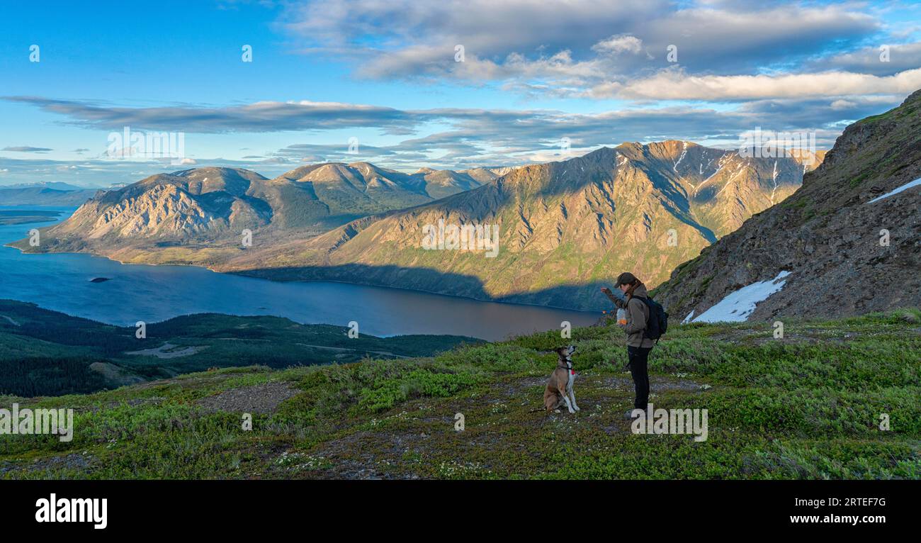 Donne in piedi su una cima di una montagna che dà da mangiare al suo cane e che si godono l'incredibile paesaggio dello Yukon lungo il Sam McGee Trail vicino a Carcross al tramonto Foto Stock
