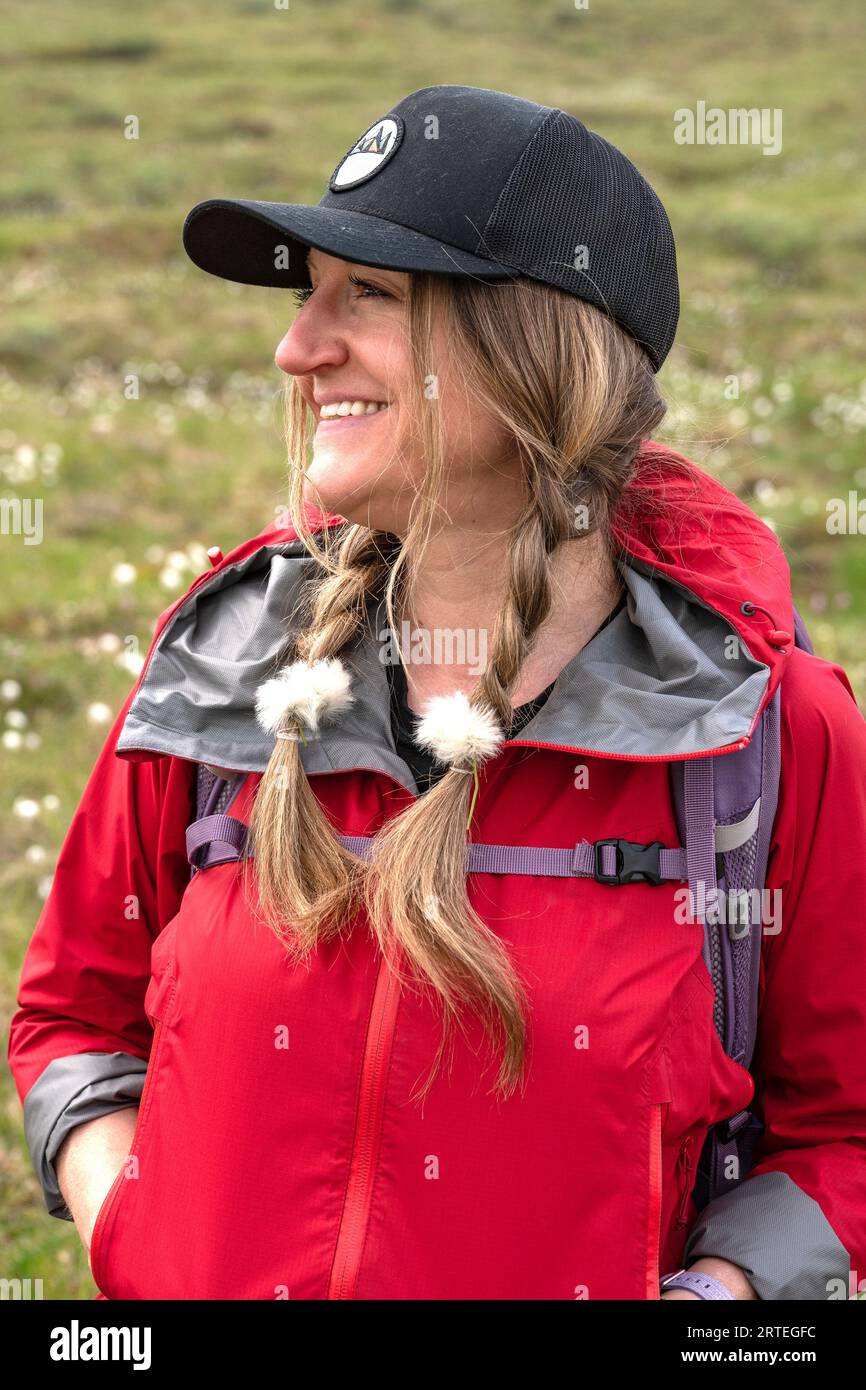 Donna sorridente con cotone artico (Eriophorum callitrix) nelle trecce dei capelli durante l'escursione; Yukon, Canada Foto Stock
