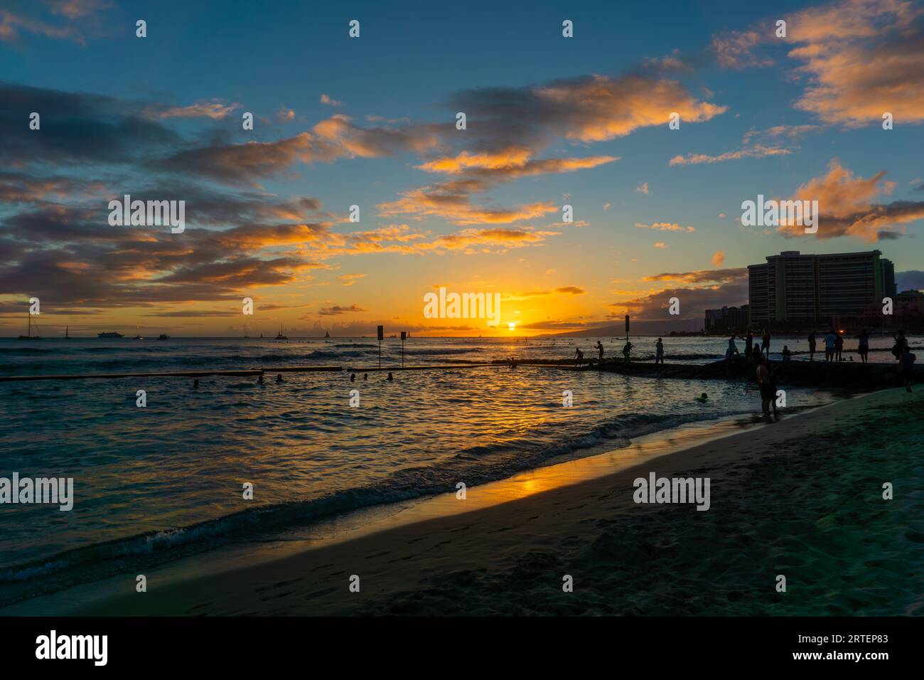 Tramonto dorato all'orizzonte sulla spiaggia di Waikiki a Oahu, Hawaii, con silhouette di visitatori non identificabili che nuotano e sulla spiaggia. Foto Stock