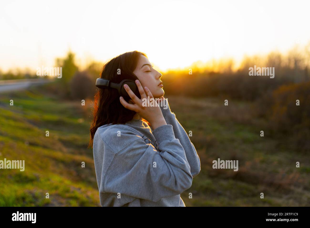 Vista laterale di una donna che ascolta musica a occhi chiusi nel prato al tramonto Foto Stock