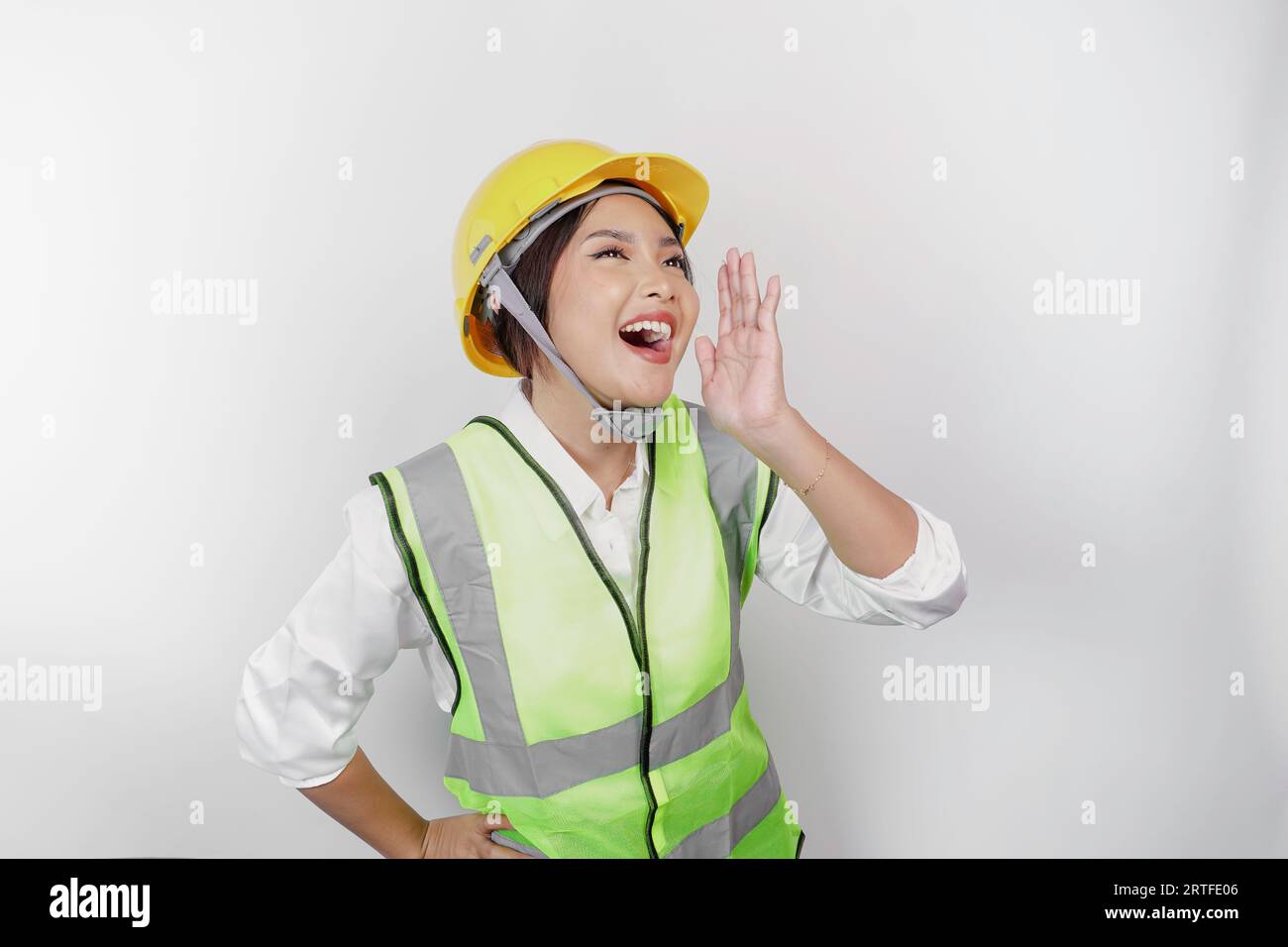 La giovane bella donna che lavora indossando un casco e un giubbotto di sicurezza grida e urla forte con una mano sulla bocca. Concetto del giorno del lavoro. Foto Stock
