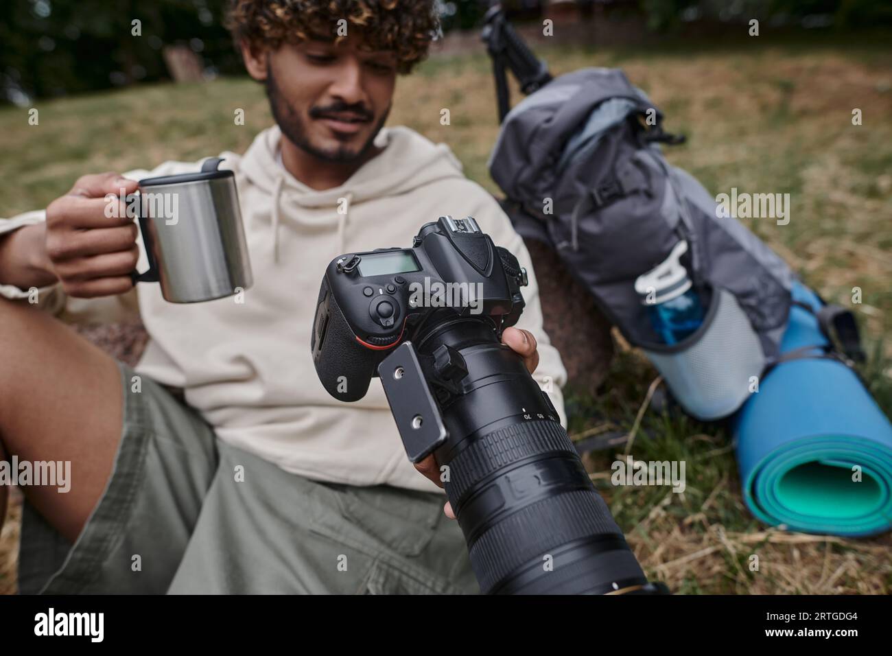 felice indiano che tiene in mano la tazza thermos e guarda le foto con la macchina fotografica, fotografo vicino all'attrezzatura da viaggio Foto Stock