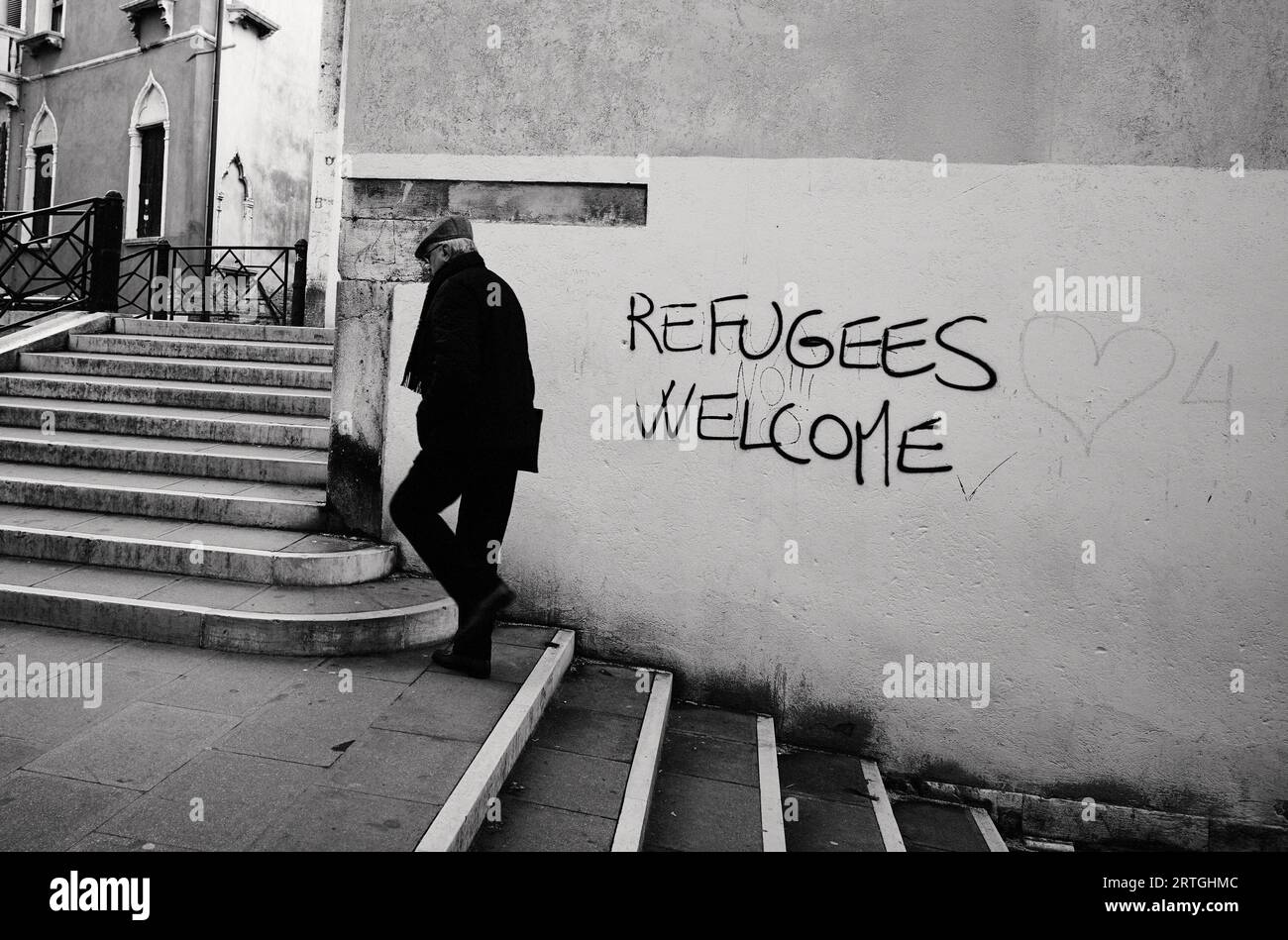 Un uomo più anziano che indossa un cappello piatto e una sciarpa che cammina fuori da un muro con graffiti. "rifugiati (no!!!!) Welcome' Venice, Italy BW Foto Stock