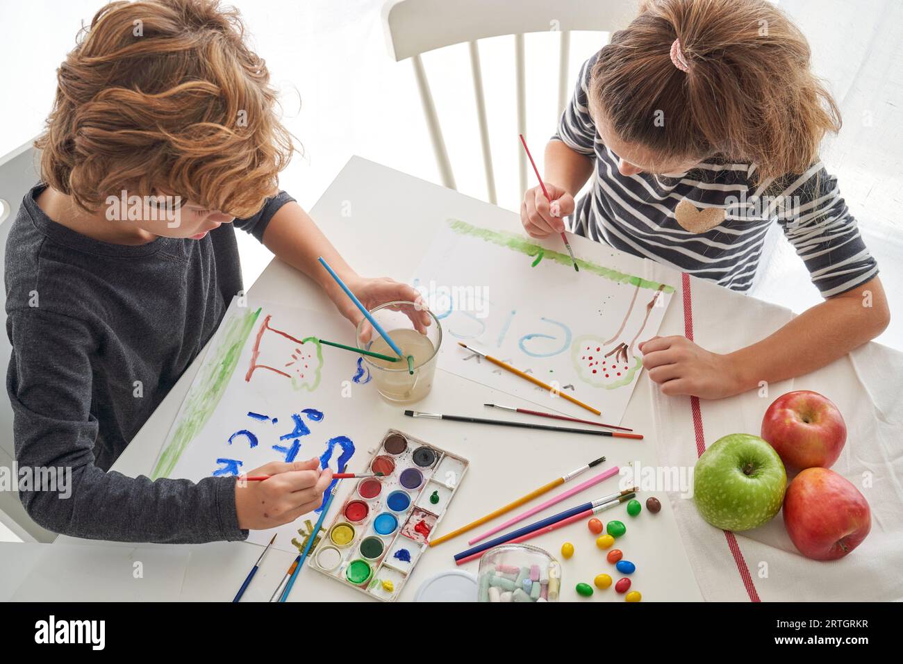 Angolo elevato di fratello e sorella che tengono pennelli che disegnano immagini su fogli di carta bianca con vernice aquarelle Foto Stock
