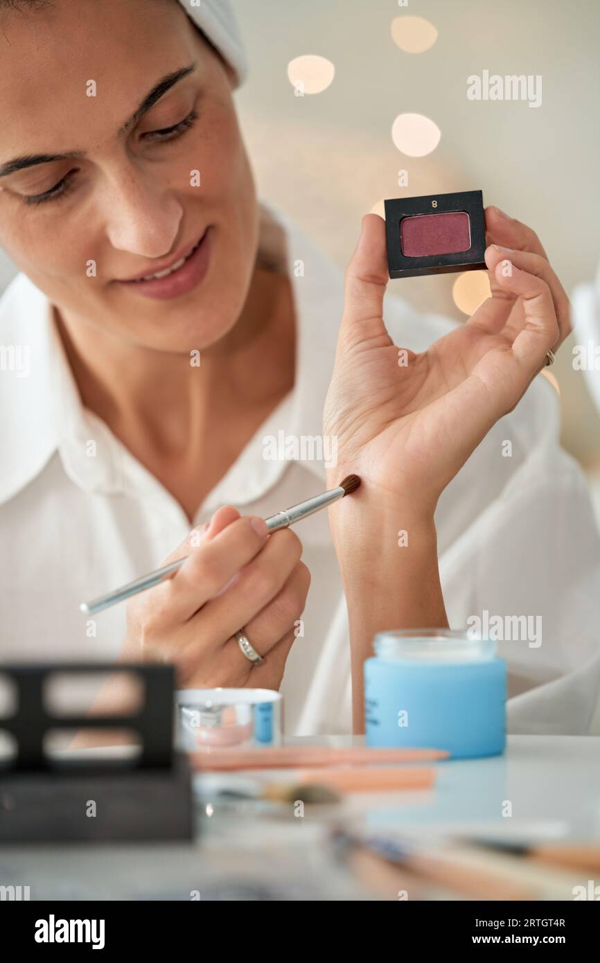 Ritaglia il visagista femminile adulto sorridente applicando un'ombra rosa sulla mano con una spazzola cosmetica durante il trucco e il test del prodotto Foto Stock
