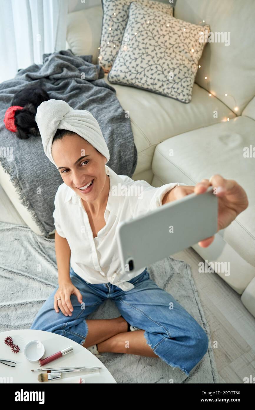 Dall'alto di una giovane blogger di bellezza con turbante per asciugamani che fa selfie su smartphone per contenuti di blog mentre si siede a terra con le croce Foto Stock