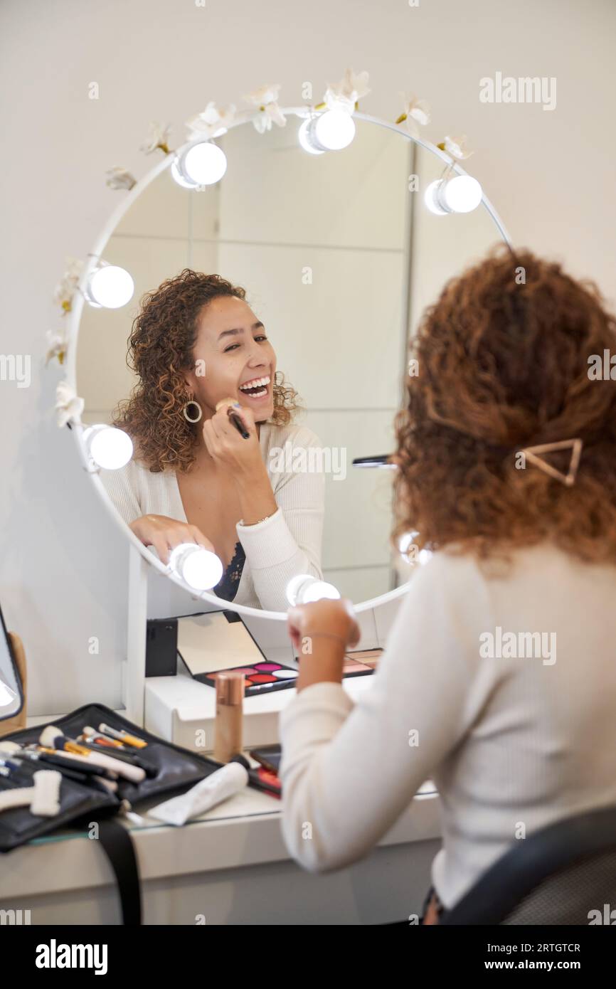 Vista posteriore di una femmina nera felice con i capelli ricci applicando la base con la spazzola mentre ridi e guarda lo specchio Foto Stock
