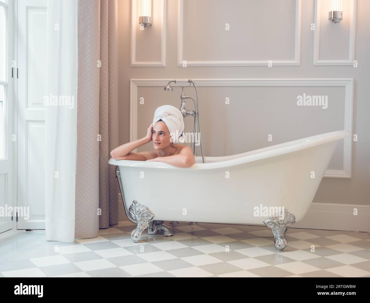 Una donna da sogno che si rilassa nella vasca da bagno, mentre si piega a mano e guarda lontano nel bagno luminoso dell'hotel Foto Stock