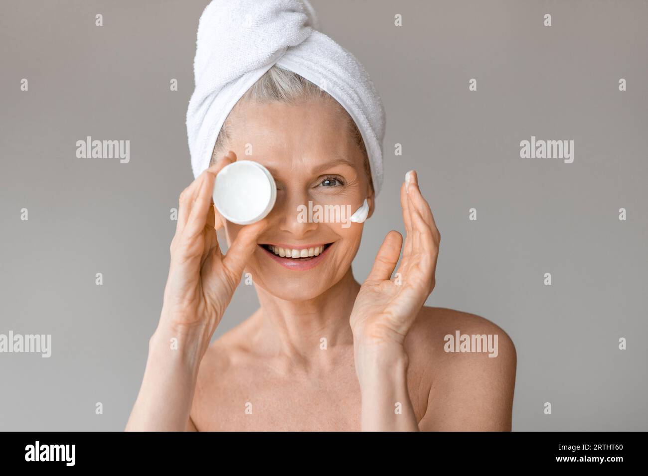 Bella donna matura che tiene in mano un vaso con crema idratante, coprendo un occhio con un prodotto per la cura della pelle Foto Stock