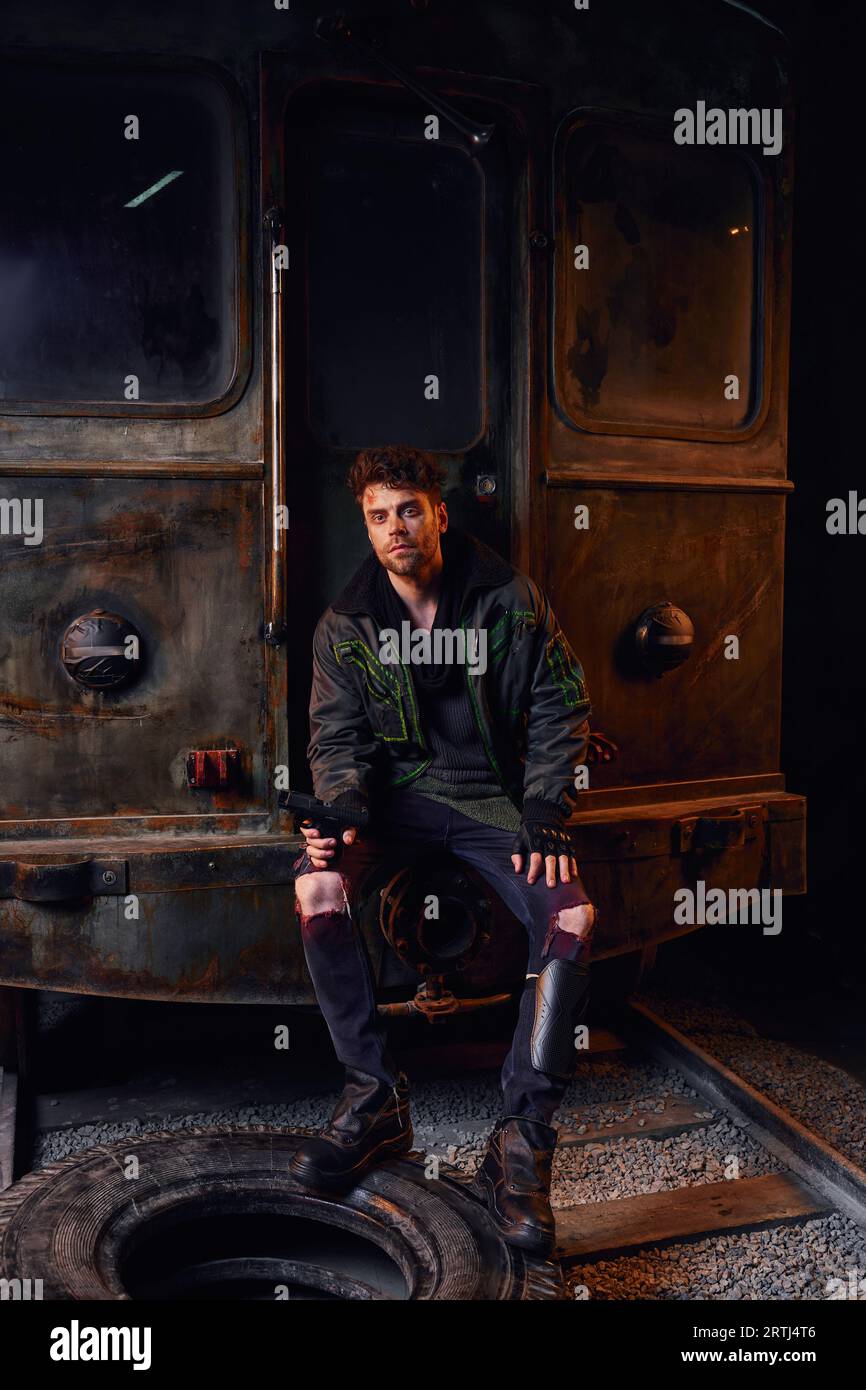 uomo non rasato con un vestito indossato seduto con la pistola vicino a carrozze arrugginite e pneumatici nella metropolitana post-disastro Foto Stock
