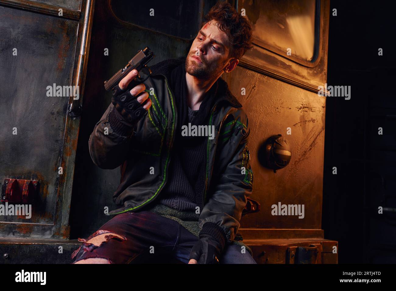 uomo non rasato in abiti indossati che guarda la pistola vicino a una carrozza arrugginita della metropolitana, sopravvivenza post-disastro Foto Stock