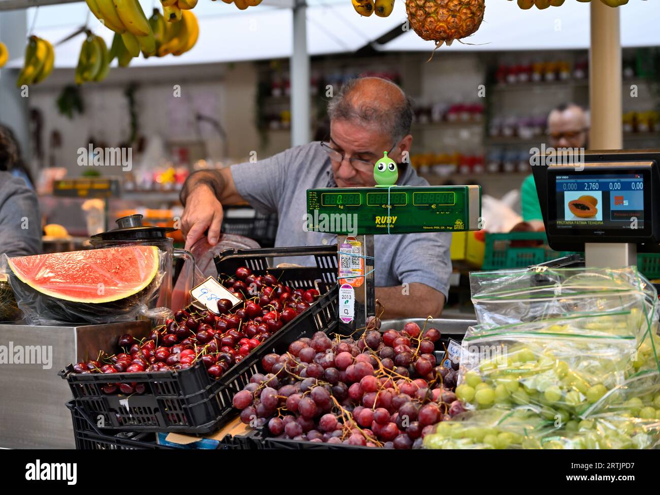 Uomo che serve al mercato con uva fresca, ciliegie e anguria in mostra con la macchina per il sollevamento pesi Foto Stock