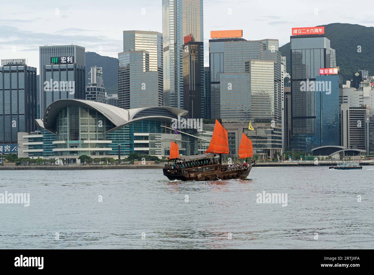 Junk boat naviga sul Victoria Harbour di giorno con il centro culturale di Hong Kong e lo skyline alle spalle. Hong Kong - 25 agosto 2023 Foto Stock