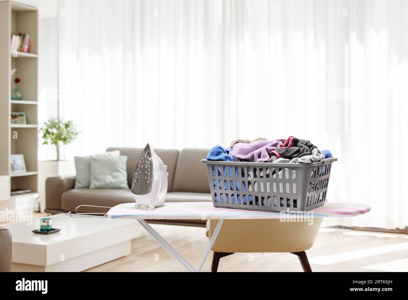 Asse da stiro con cestino per la lavanderia pieno di vestiti all'interno del soggiorno Foto Stock