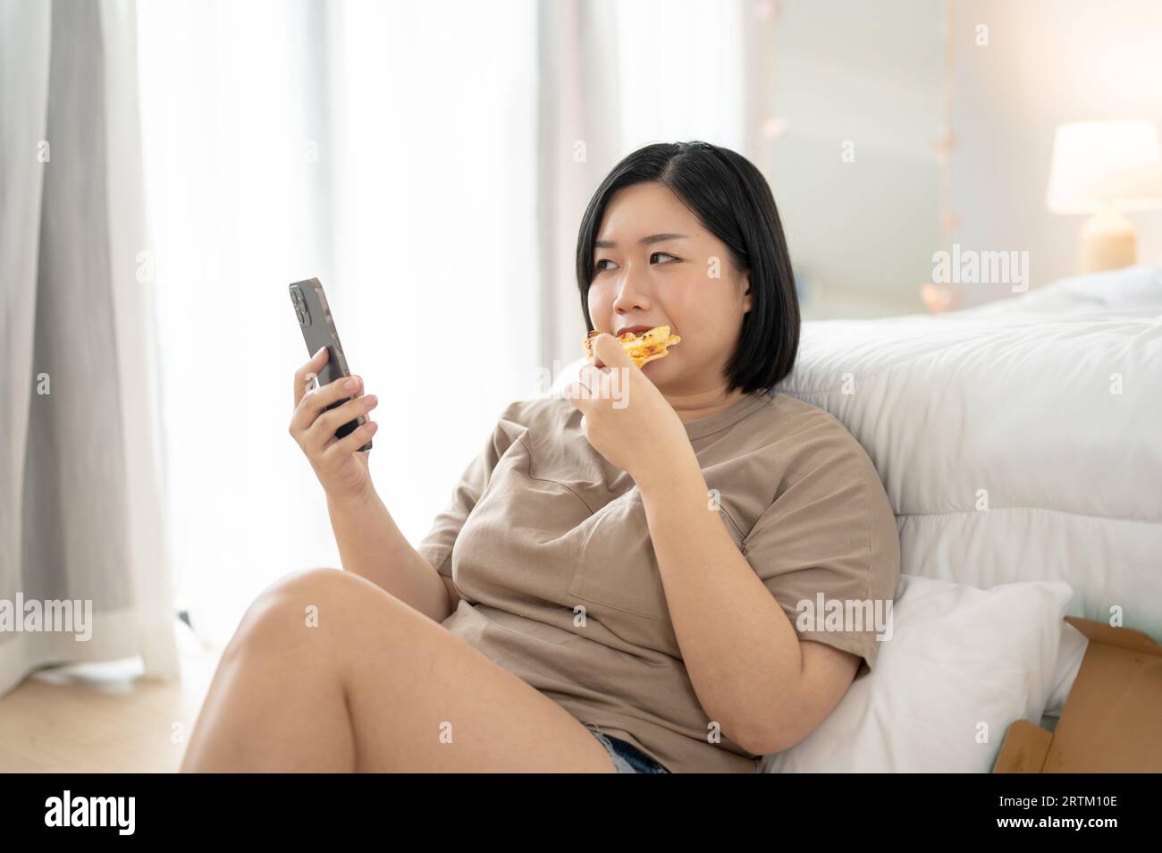 Una donna asiatica curvy Plus-size, felice e gioiosa, in abiti comodi, si sta gustando la pizza mentre scorre i social media sul suo smartphone nel suo bedroo Foto Stock