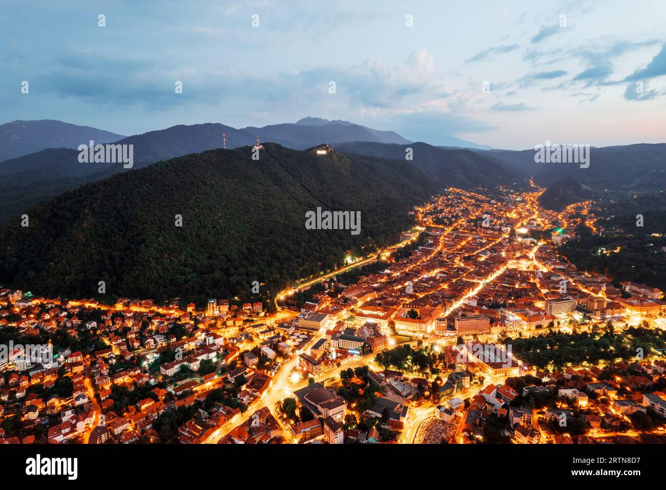 Vista aerea di Brasov al tramonto, Romania. Centro storico con edifici antichi e illuminazione, colline ricoperte di verde Foto Stock