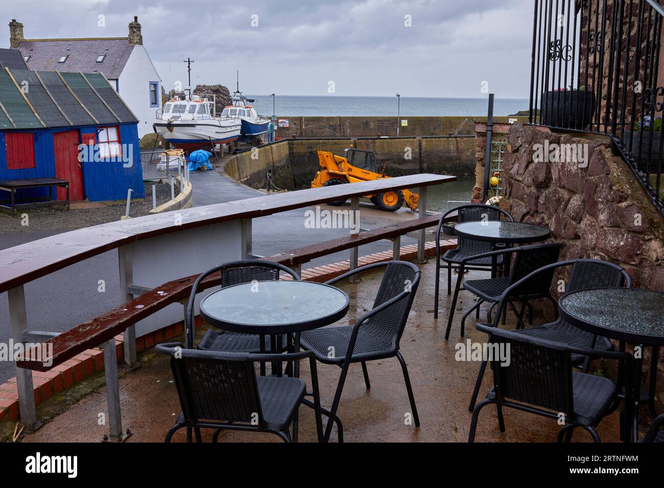 Davanti al tavolino bagnato e vuoto, si può ammirare la vista del porto di St Abbs, con le barche da pesca appoggiate sul muro del porto per lavori di manutenzione Foto Stock