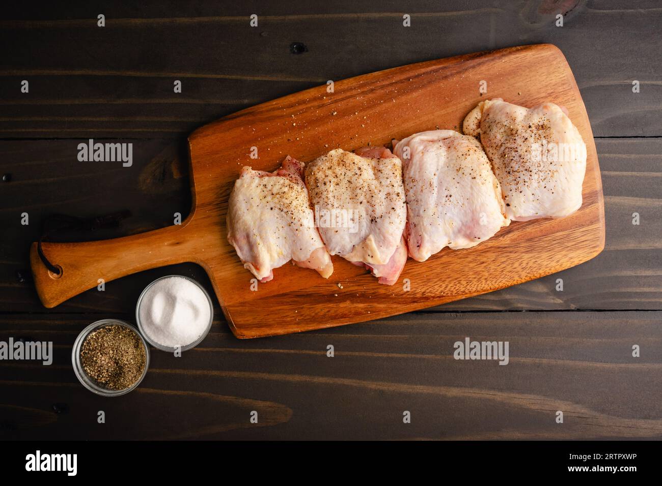 Cosce di pollo crudo condite con buccia su un tagliere di legno: Tagli crudi di di pollame di carne scura conditi con sale marino e pepe nero Foto Stock