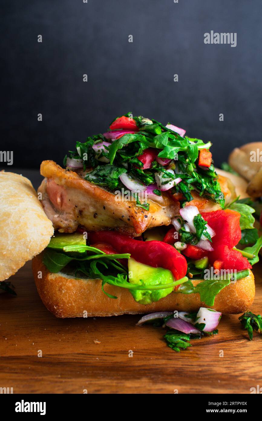 Sandwich di pollo Chimichurri su tagliere di legno: Panino aperto con avocado, peperoni arrostiti e rucola Foto Stock