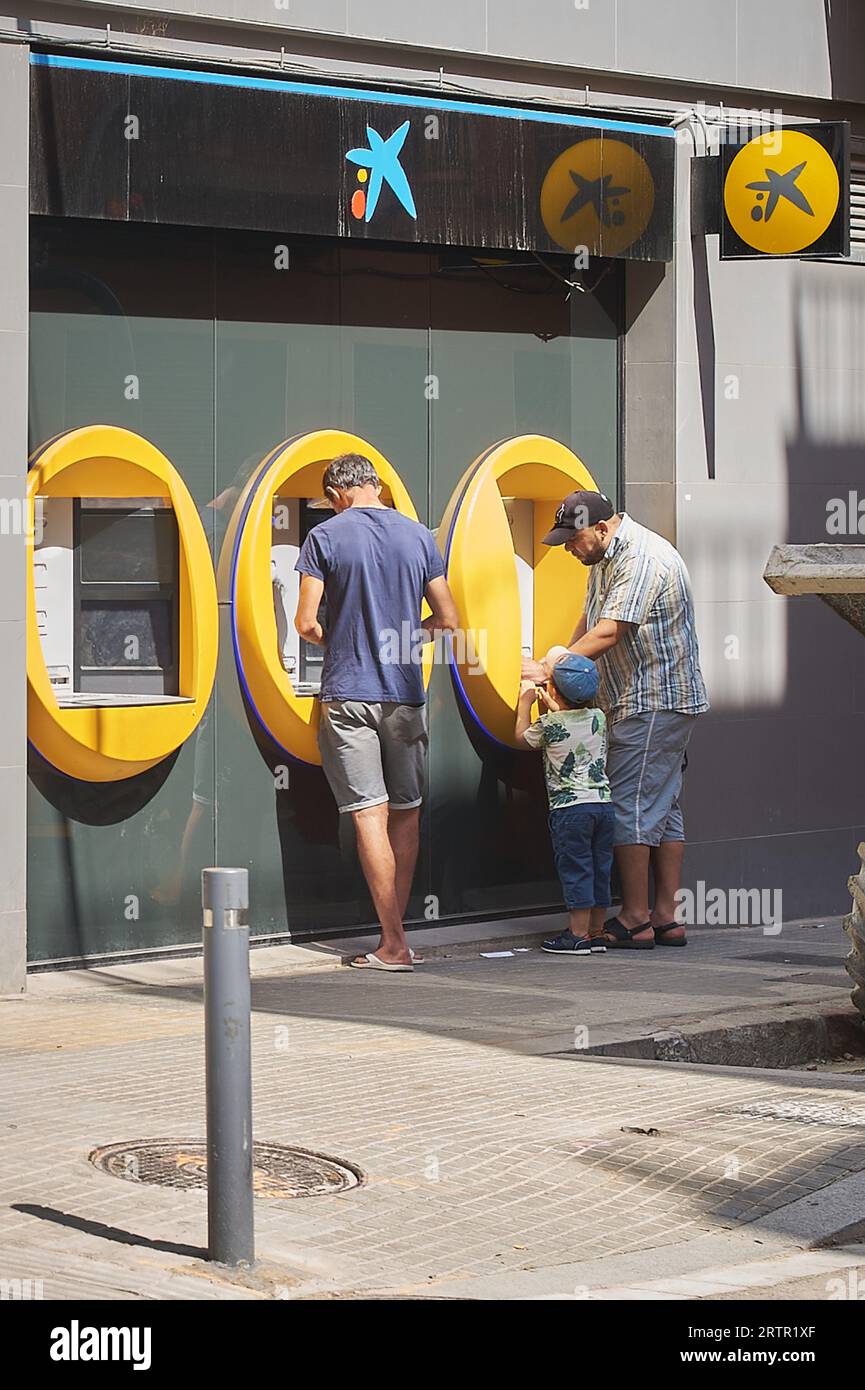 Viladecans, Spagna - 14 settembre 2023: Due uomini e un bambino davanti al bancomat Caixa ritirano denaro in una giornata di sole. Foto Stock