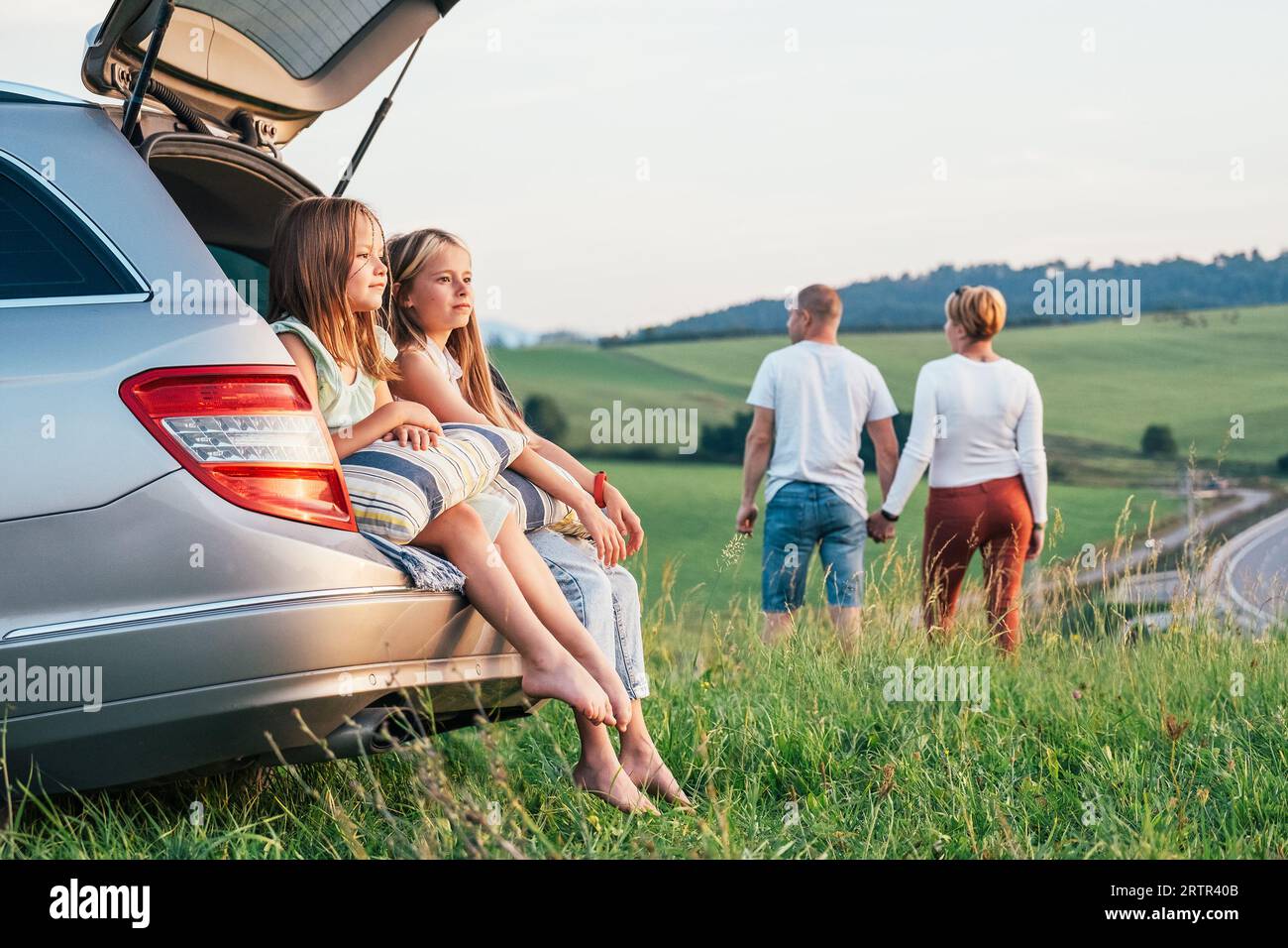 Due sorelle che si trovano sedute nel bagagliaio dell'auto e si godono la luce del tramonto. Giovani coppie caucasiche hanno una pausa auto con mano in mano camminare vicino a prati verdi Foto Stock