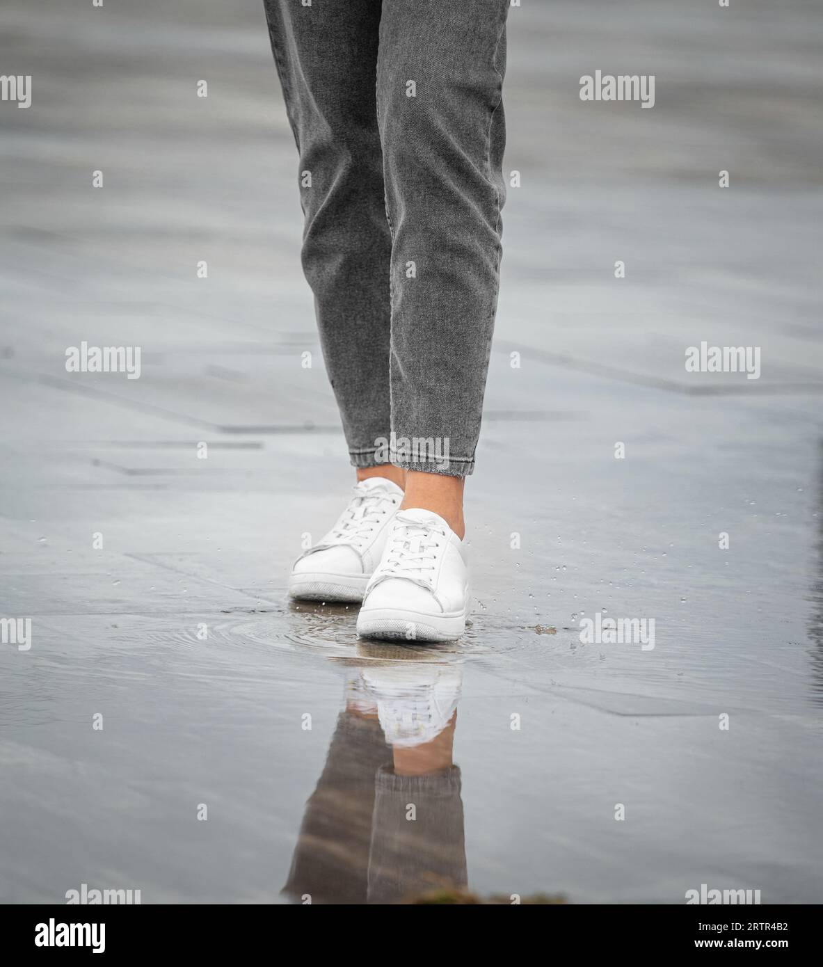 Le gambe di una ragazza in jeans e scarpe da ginnastica bianche camminano in una strada della città con pioggia, gocce di pioggia, spruzzi d'acqua, riflessi in pozzanghere Foto Stock