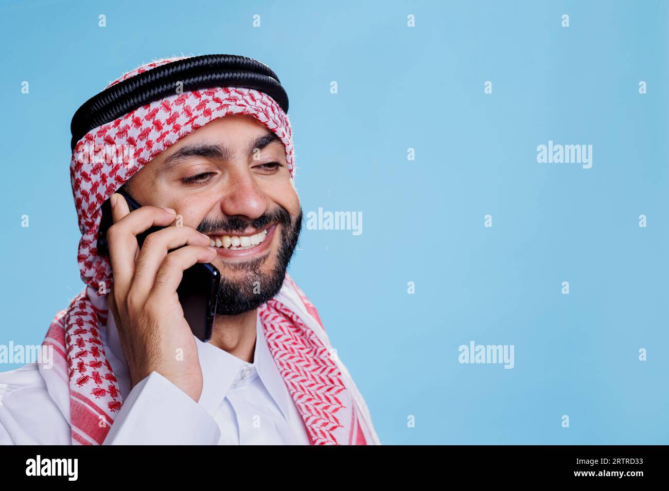 Uomo musulmano sorridente che indossa un velo ghutra con fascia di corda che parla al telefono cellulare. Allegro arabo vestito di copricapo islamico che parla su smartphone con felice espressione Foto Stock