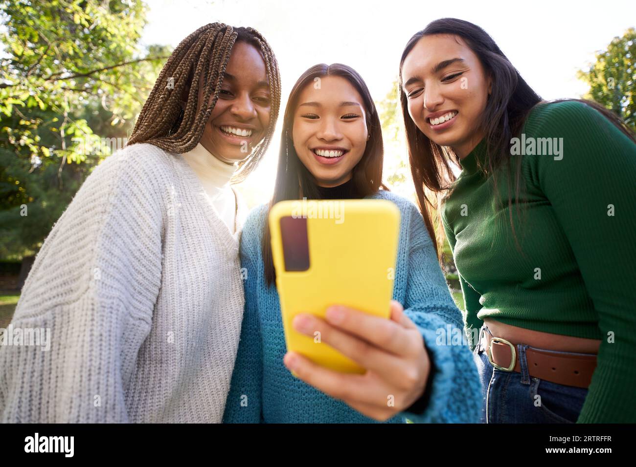Tre ragazze sorridenti e multirazziali che utilizzano il parco all'aperto con telefoni cellulari. Persone felici connesse online. Foto Stock