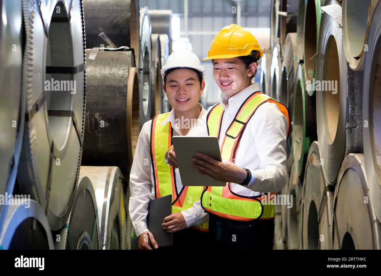 Due operai dedicati, adornati con elmetti protettivi, si trovano in posizione sicura nell'arena industriale, circondata da enormi bobine di acciaio. Foto Stock